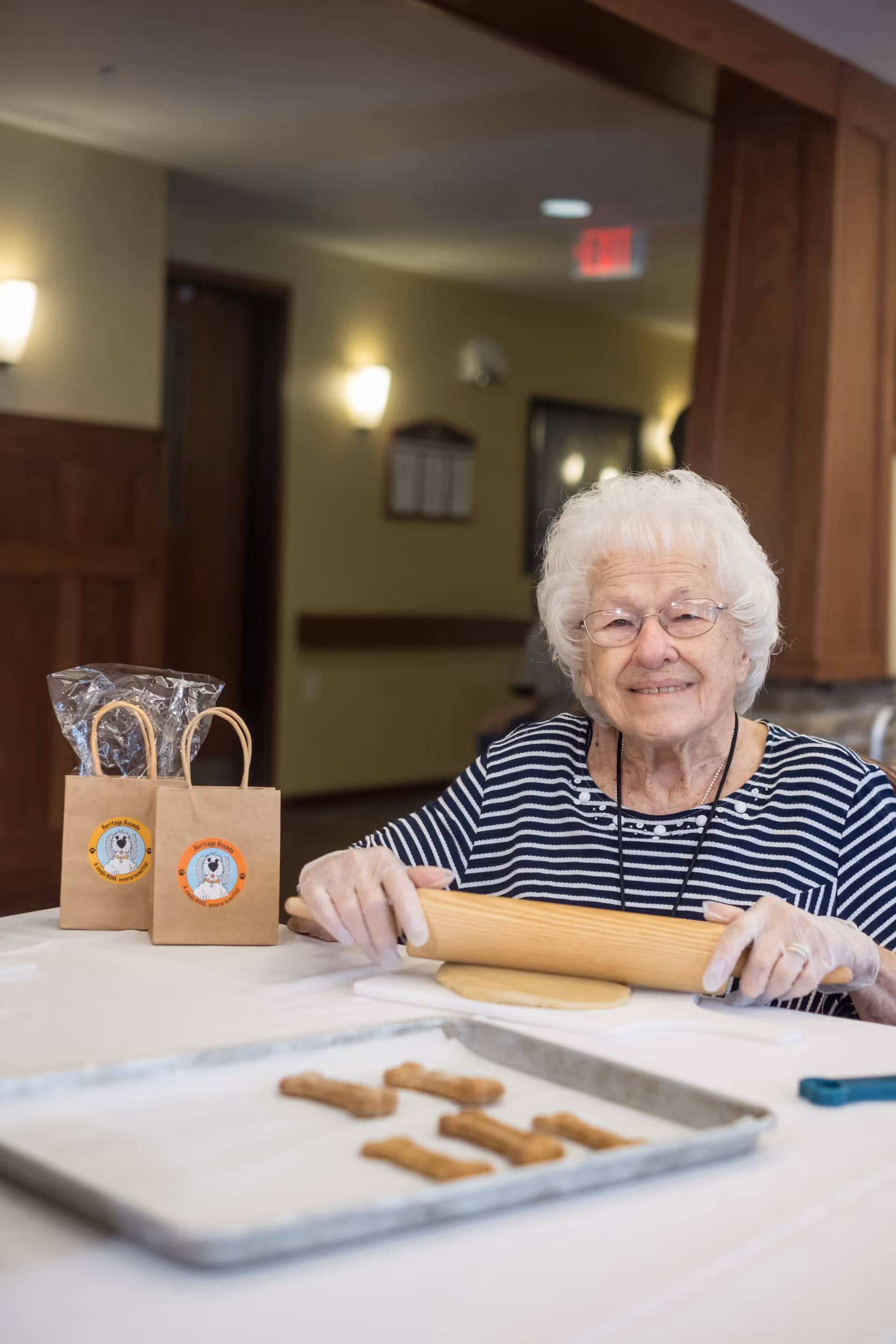 An elderly woman with white hair and glasses is smiling while rolling out dough with a wooden rolling pin at a table. In front of her is a baking tray with bone-shaped cookies. Two small brown paper bags with dog-themed stickers are also on the table. The setting appears to be a cozy indoor area with warm lighting.
