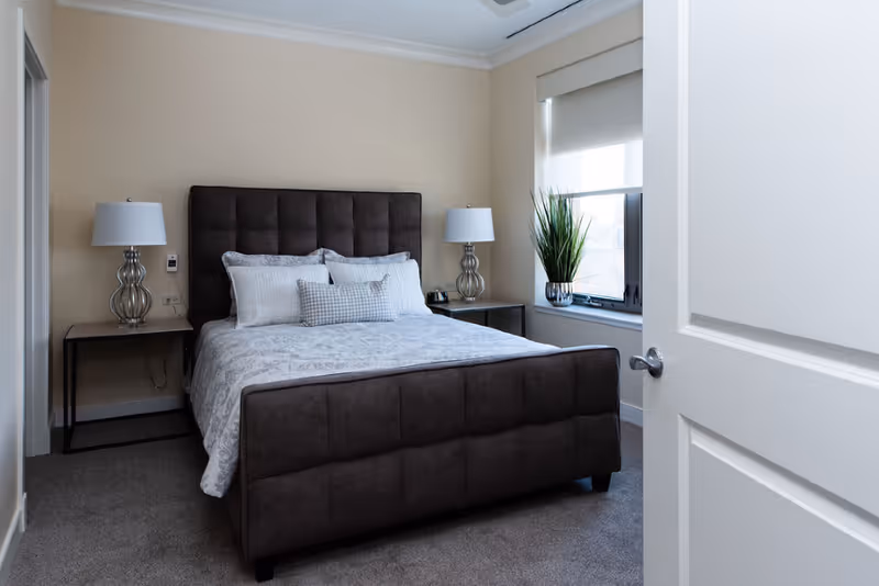 Neatly made bedroom with a tufted dark upholstered bed, matching bedside tables and lamps, and a window with a potted plant.