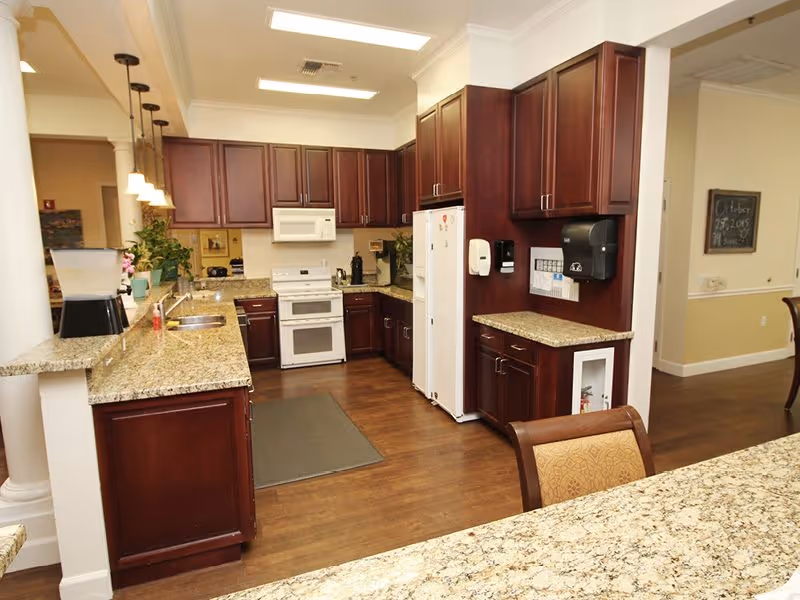 A spacious kitchen area with dark wood cabinets, granite countertops, a white stove, microwave, and refrigerator. The kitchen has a wooden floor and is well-lit with ceiling lights and pendant lights over the counter. There is a water dispenser on the counter and a paper towel dispenser on the wall. Adjacent to the kitchen is a dining area with a chair partially visible.