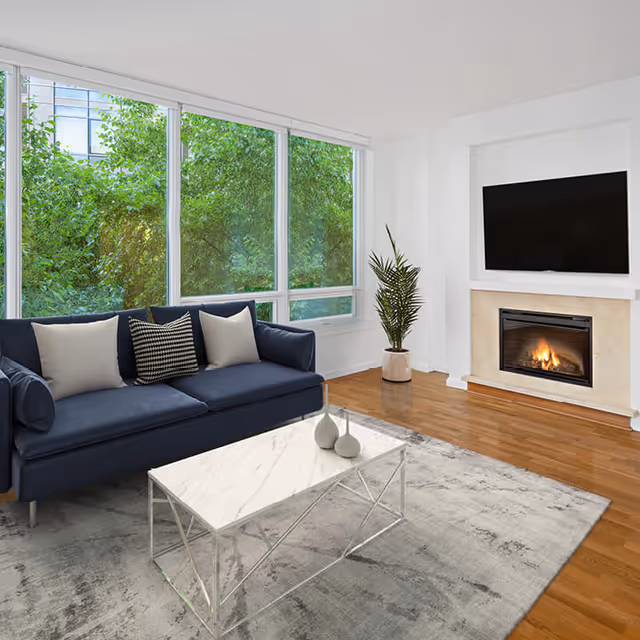 A modern living room with a dark blue sofa adorned with three cushions, a white marble coffee table with two decorative vases, a large window showing green foliage outside, a potted plant in the corner, a fireplace with a fire burning, and a flat-screen TV mounted above the fireplace.