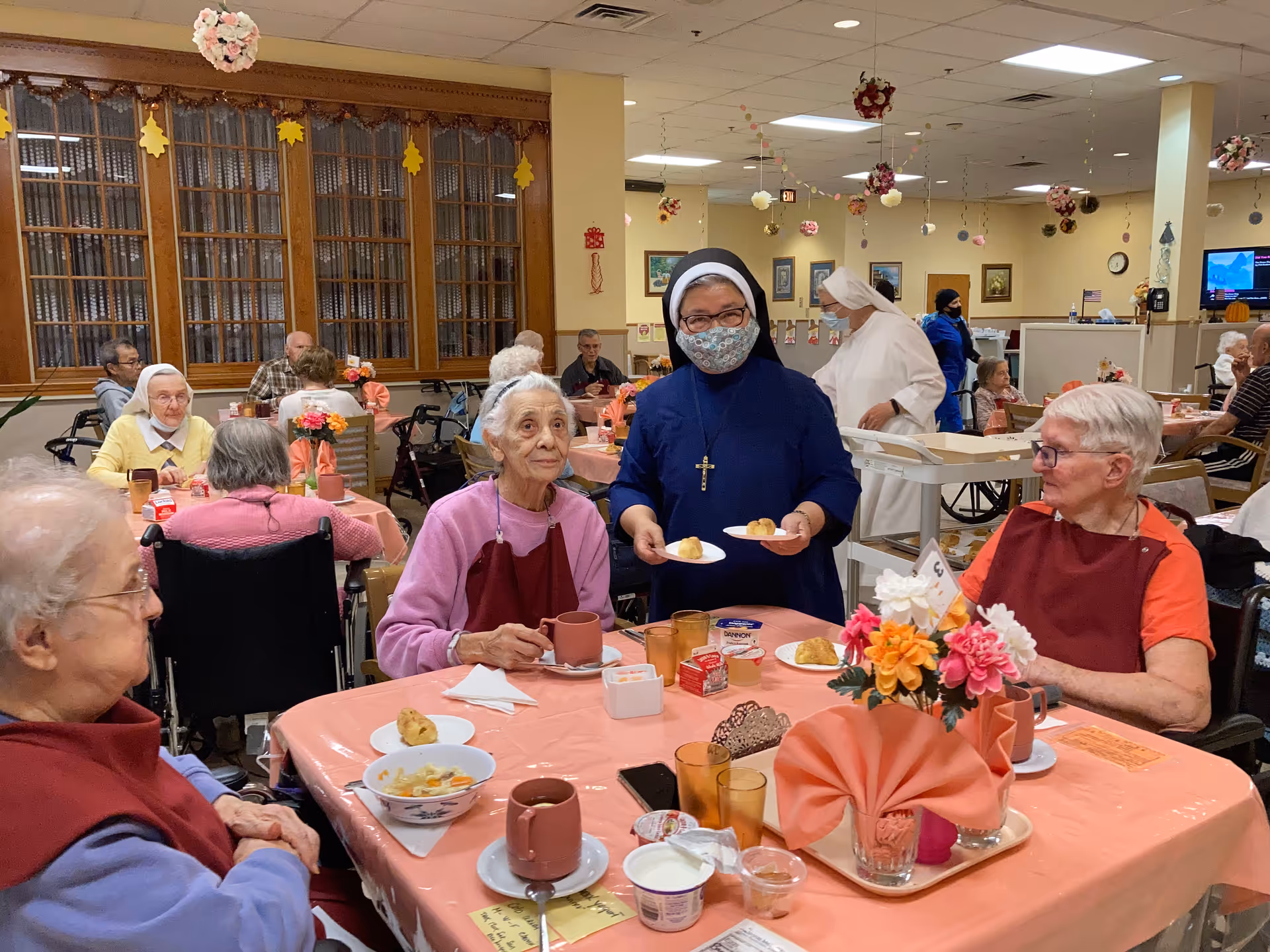 A dining room in a senior living facility where elderly residents are seated at tables covered with pink tablecloths, enjoying a meal. A nun wearing a mask and a cross necklace is serving food to the residents. The room is decorated with hanging flowers and autumn-themed decorations, and other staff and residents are visible in the background.
