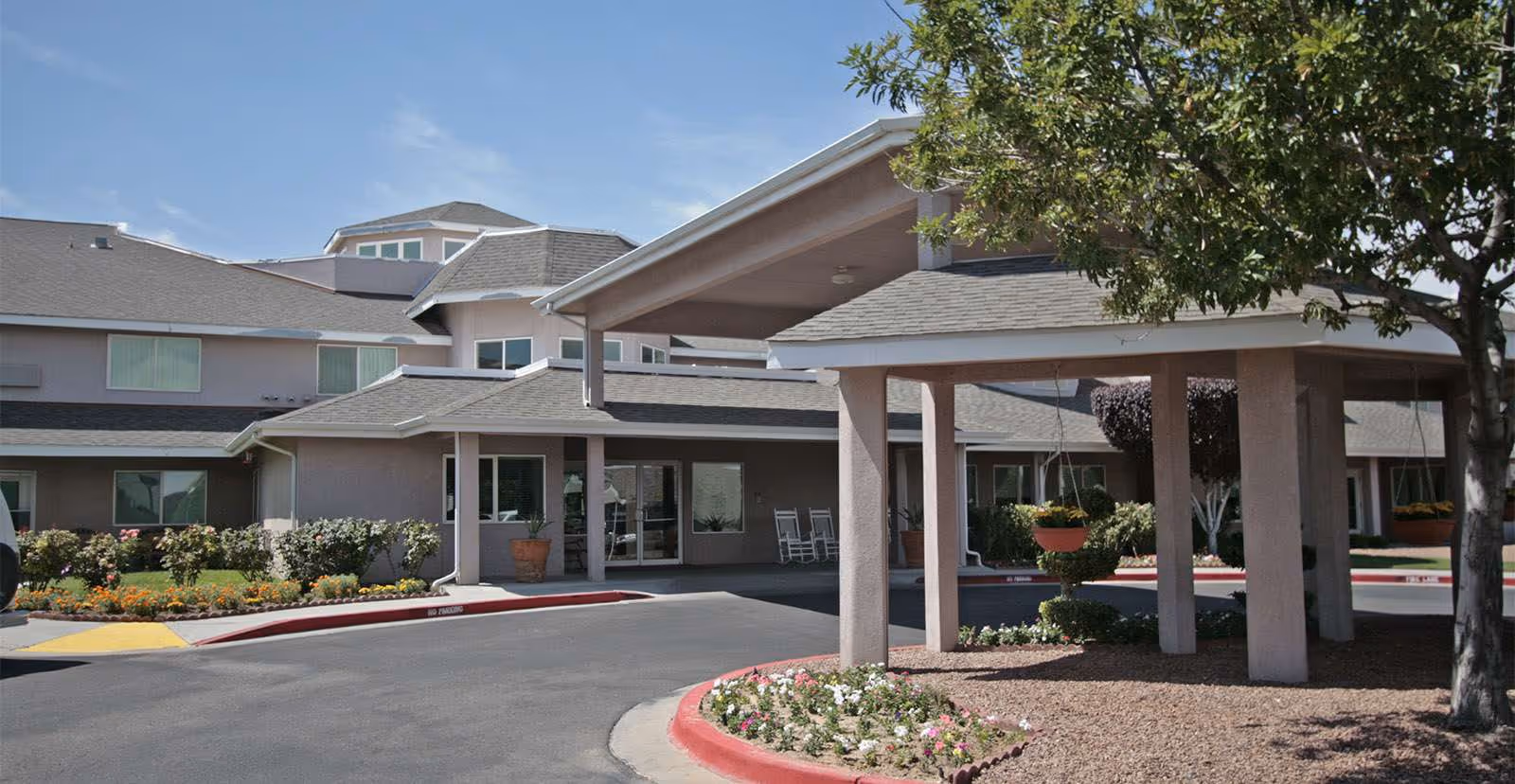 Exterior view of Solstice Senior Living at Rio Norte showing the entrance with a covered driveway, landscaped flower beds, and a tree on the right side.