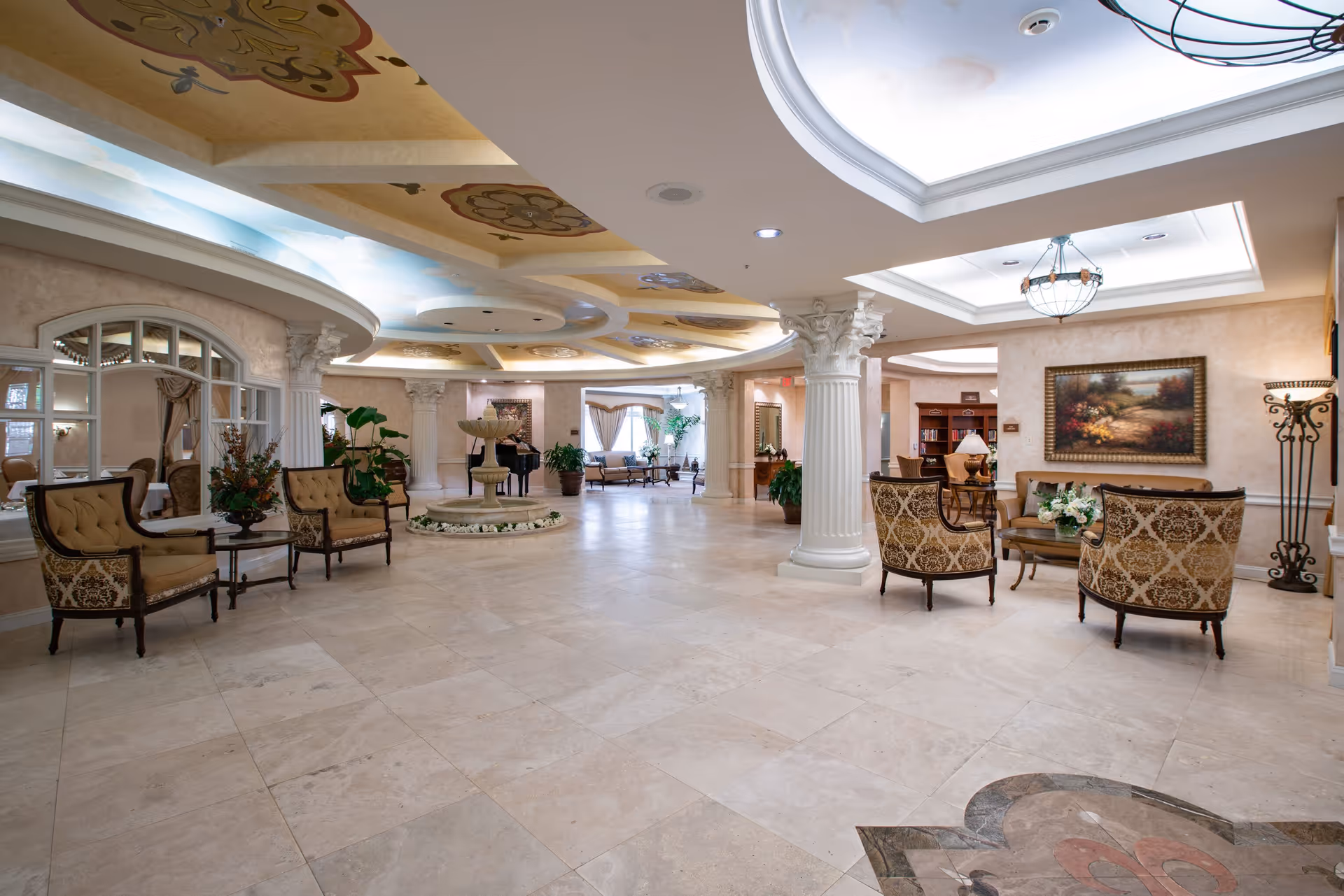 Spacious and elegant senior living facility common area with marble flooring, ornate ceiling designs, classical columns, and comfortable seating arrangements including armchairs and sofas. A decorative fountain is centrally placed, and the room is well-lit with natural light from windows and ceiling fixtures. There are plants and framed artwork enhancing the warm and inviting atmosphere.