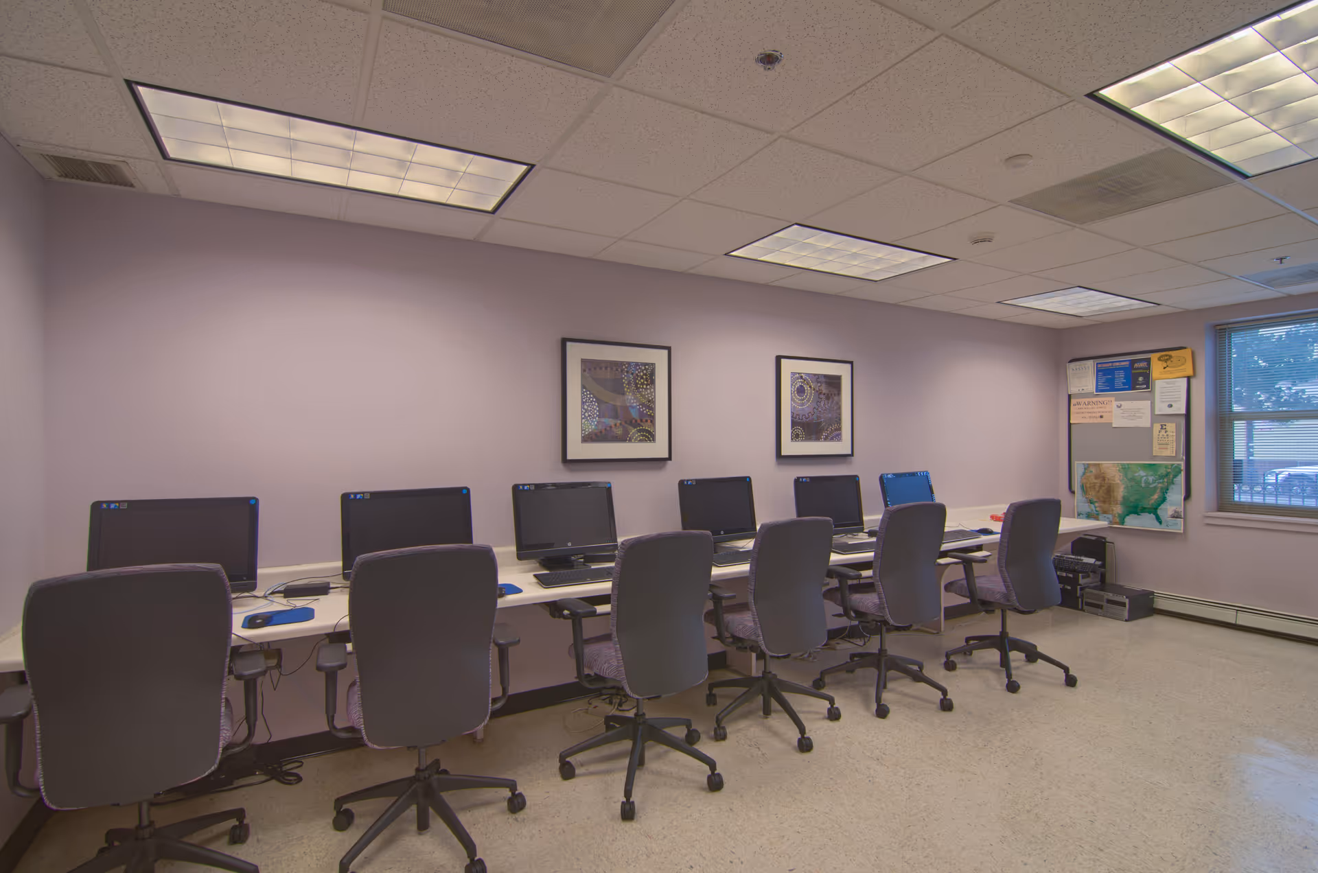 A computer room with a row of seven desktop computers on a long white desk. Each computer has a black office chair in front of it. The walls are light purple with two framed abstract art pieces hanging above the computers. There is a window on the right side with a view outside and a bulletin board with various notices and a map below it. The ceiling has fluorescent lights.