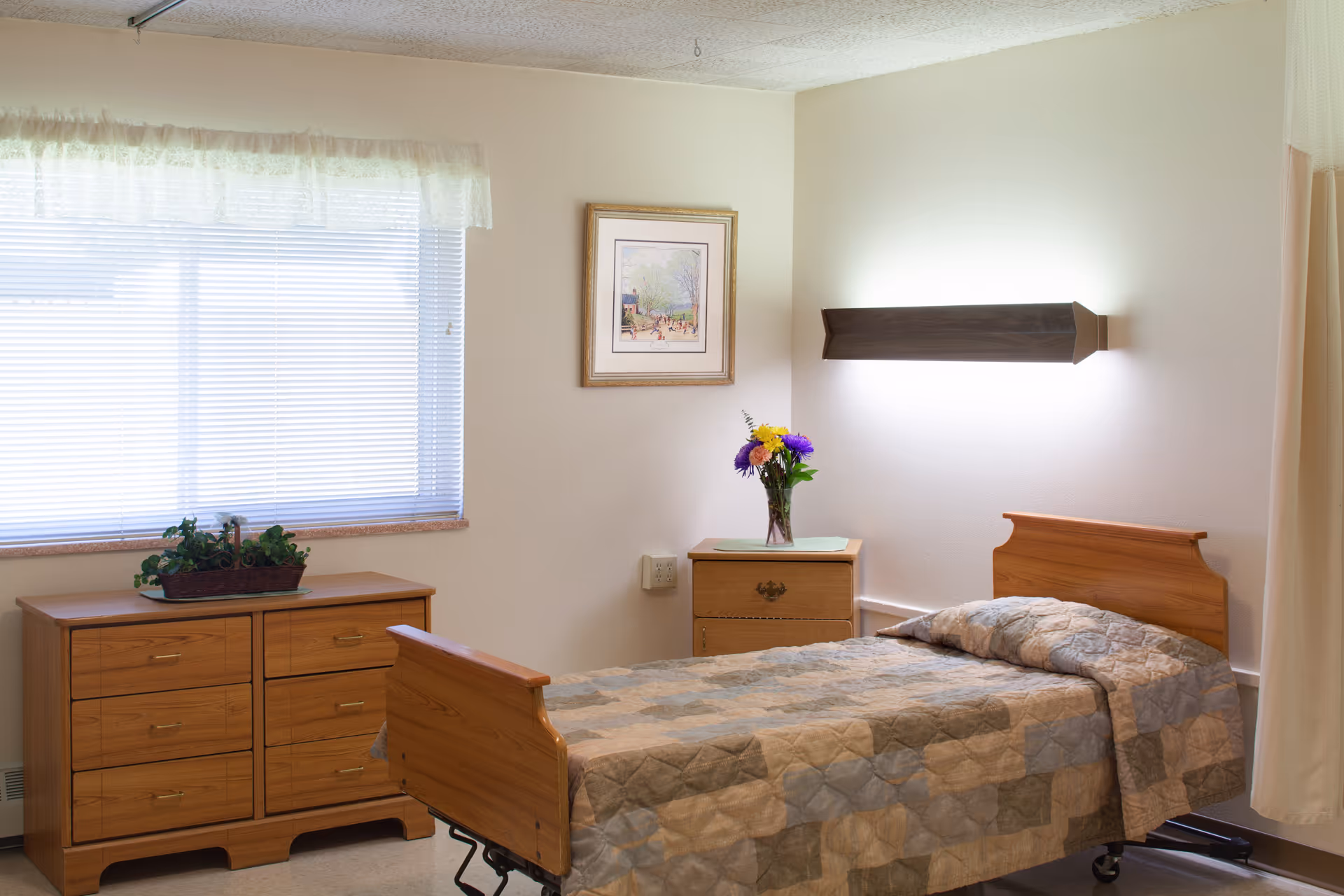Simple senior resident bedroom with a single wooden bed, quilted blanket, dresser, nightstand with flowers, and a window with blinds.
