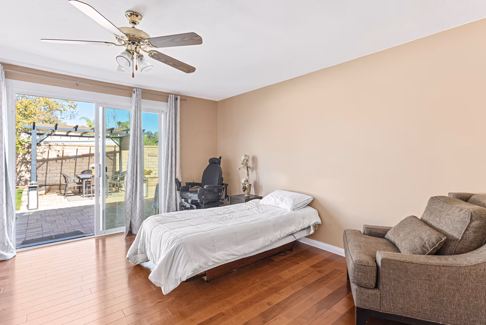 A bedroom with a single bed covered in a white comforter, a brown armchair with a matching pillow, a ceiling fan with lights, and a sliding glass door leading to an outdoor patio with a table and chairs. There is also a black wheelchair next to the bed and a small nightstand with a vase of flowers.