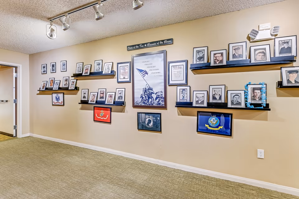 Interior hallway wall decorated with framed photographs and military memorabilia, including a central poster of the Iwo Jima flag raising and various portraits of servicemen and women displayed on shelves and mounted on the wall.