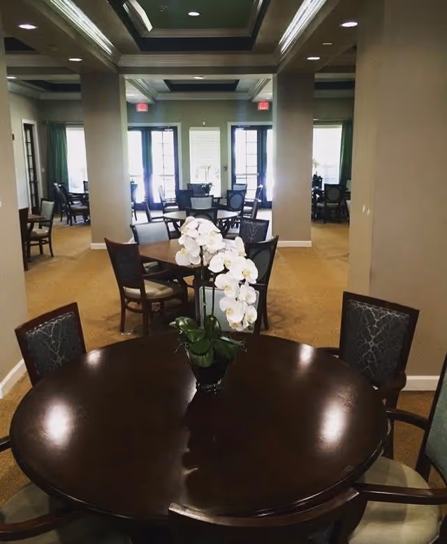 Interior view of a senior living facility dining room with round wooden tables and chairs. A white orchid centerpiece is on the closest table. The room has beige carpet, columns, and large windows with glass doors letting in natural light.
