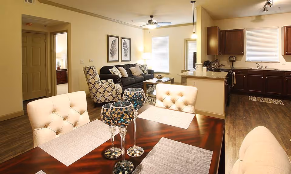 Open-plan living and dining area with a wooden dining table and tufted chairs in the foreground, a sofa and patterned accent chair in the middle, and a kitchen with dark cabinets in the background.