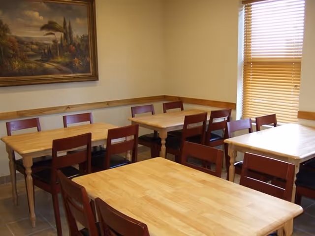 A small dining room with several wooden tables and matching wooden chairs with dark seats. There is a window with closed blinds on the right wall and a framed landscape painting on the left wall.