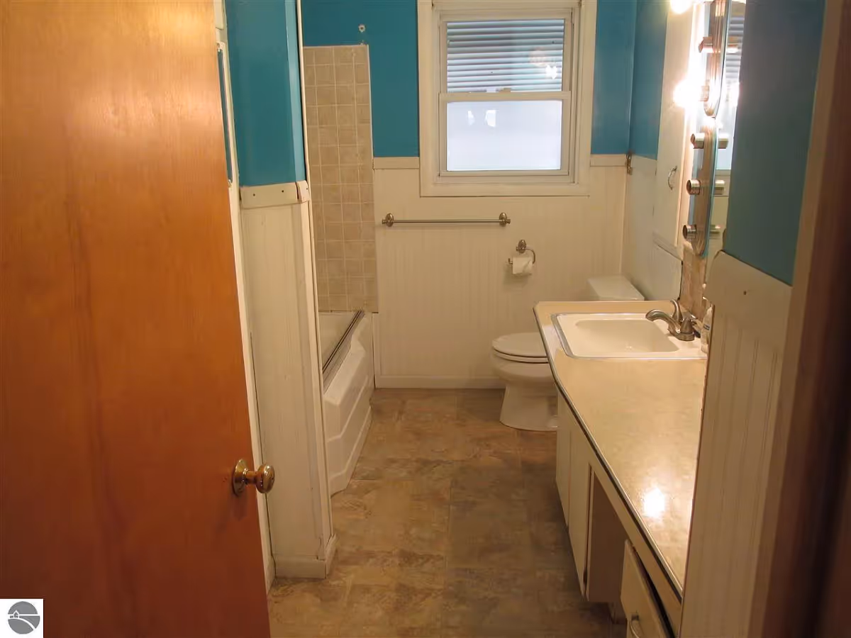 Interior view of a bathroom with a wooden door partially open, a bathtub with tiled wall, a toilet, a sink with countertop, and a window with blinds. The walls are painted blue above white wainscoting, and the floor has beige tiles.