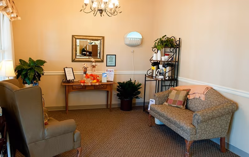A cozy sitting area in a senior living facility with a patterned loveseat adorned with a plaid pillow and a pink throw blanket, a cushioned armchair, a small wooden table with decorative items and a mirror above it, a metal shelving unit holding coffee supplies and plants, and a wall-mounted light fixture. The room has beige walls, carpeted floor, and soft lighting from a chandelier and a table lamp.