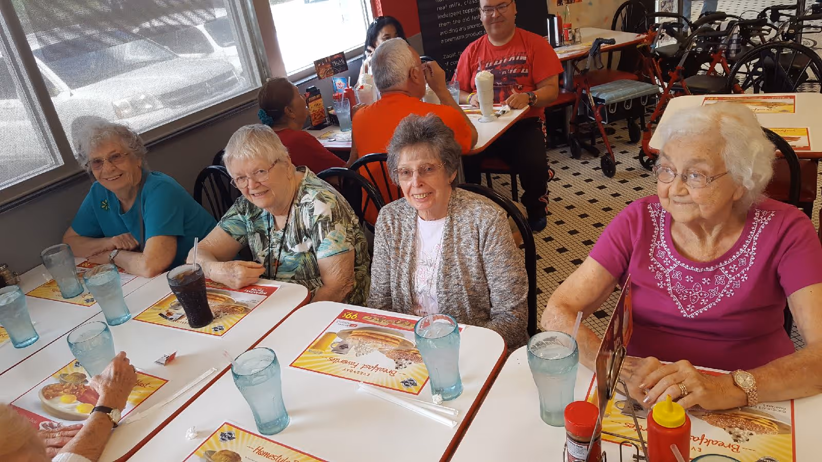 A group of elderly women sitting around a table in a dining area, smiling and enjoying each other's company. The table has placemats, glasses of water, and a glass of soda. In the background, other people are seated at tables, and there are walkers visible near the wall.