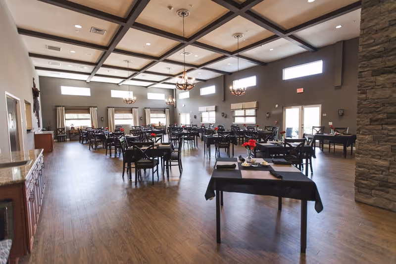 A spacious dining room with multiple tables and chairs arranged neatly. Each table is set with black tablecloths, napkins, and small flower vases with red flowers. The room has large windows with beige curtains, wooden flooring, and a high ceiling with decorative beams and hanging chandeliers. There is a stone wall on the right side and a cabinet with a granite countertop on the left.