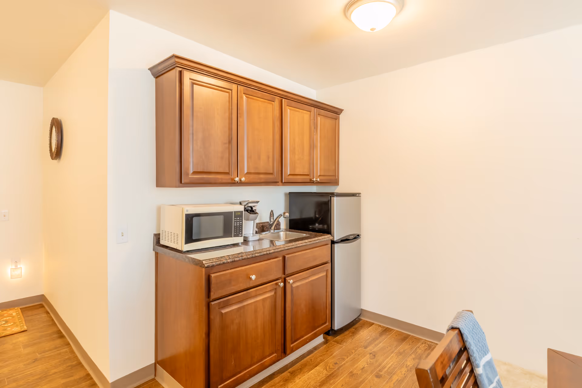 A small kitchenette area with wooden cabinets, a countertop with a microwave, a coffee maker, and a small sink. Next to the counter is a compact refrigerator. The floor is wooden, and a chair with a blue towel draped over it is partially visible on the right side.