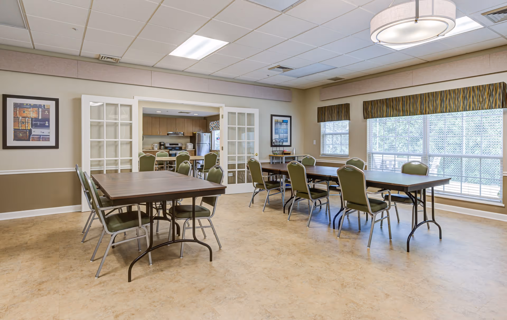 A bright dining room with several rectangular tables and green chairs arranged around them. The room has large windows with patterned valances letting in natural light. There are framed pictures on the walls and a modern circular ceiling light fixture. In the background, a kitchen area is visible through open double glass doors.