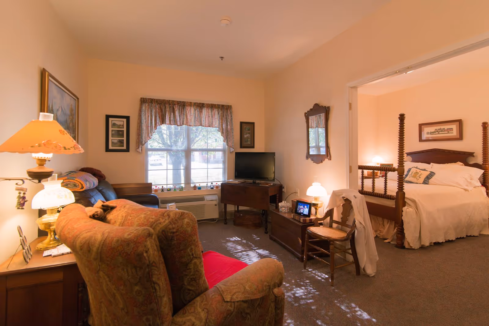 A cozy living room area with a patterned armchair, a wooden side table with a lamp, a leather couch with a colorful blanket, and a window with floral curtains. A small TV sits on a wooden table against the wall. To the right, an open doorway leads to a bedroom with a wooden four-poster bed, white bedding, a wooden chair with a white cloth draped over it, and a bedside table with a lamp.