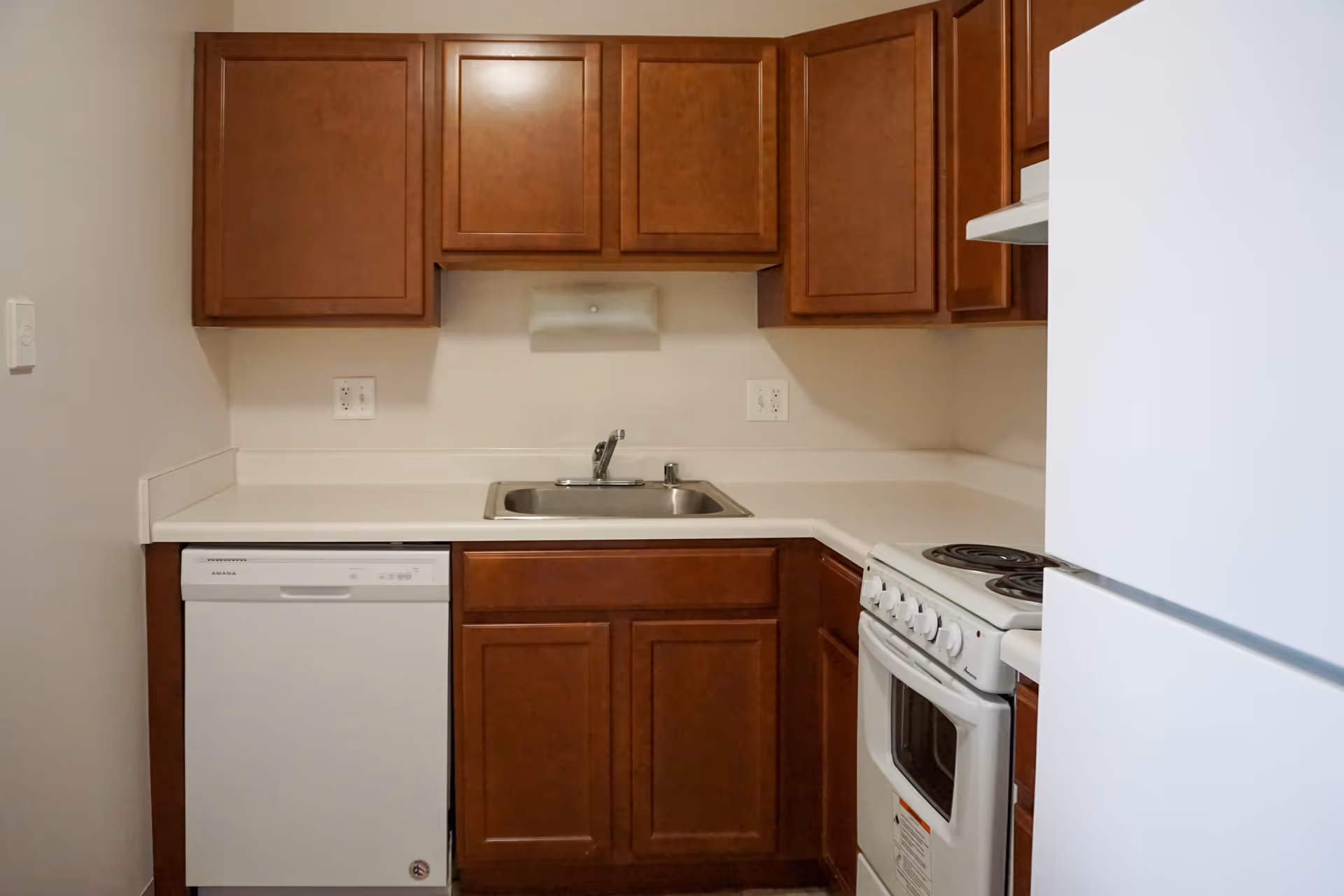 Small kitchen with brown wooden cabinets, a white countertop, a stainless steel sink, a white dishwasher, a white electric stove with four burners, and a white refrigerator.