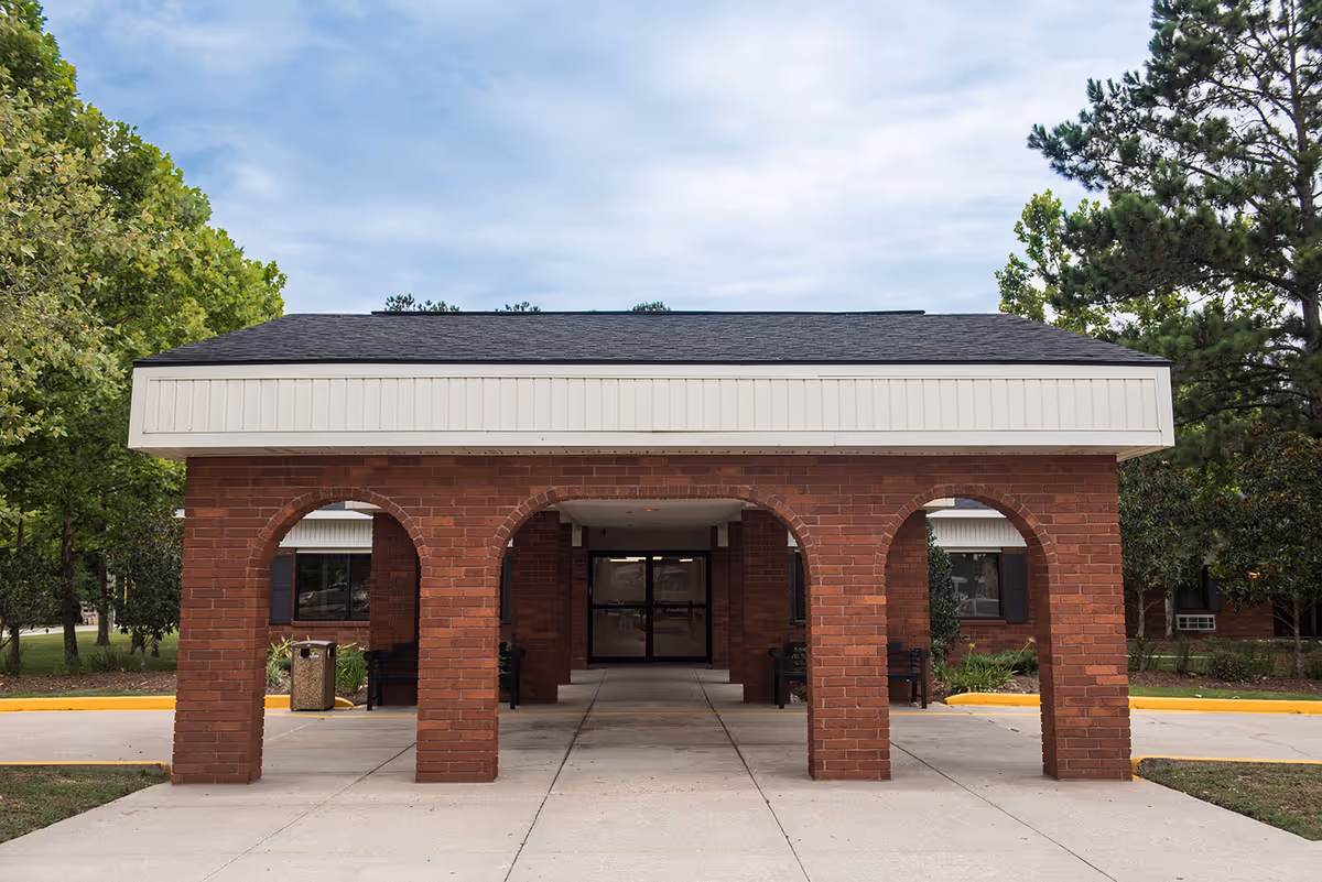 Entrance of a brick building with a covered walkway supported by four brick arches, surrounded by trees and greenery under a partly cloudy sky.