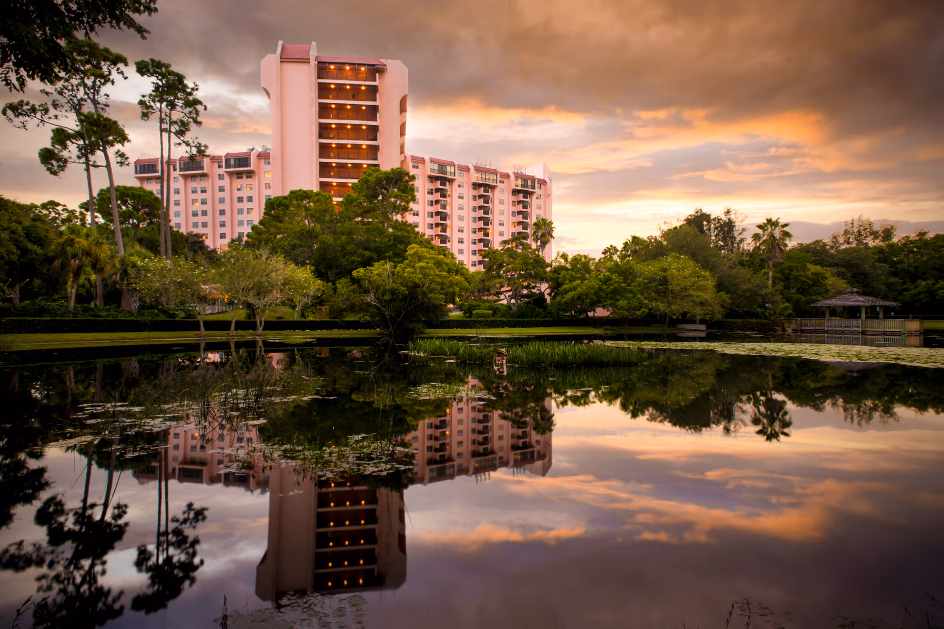 A tall pink residential building with balconies is reflected in a calm pond surrounded by trees and greenery during a colorful sunset sky.
