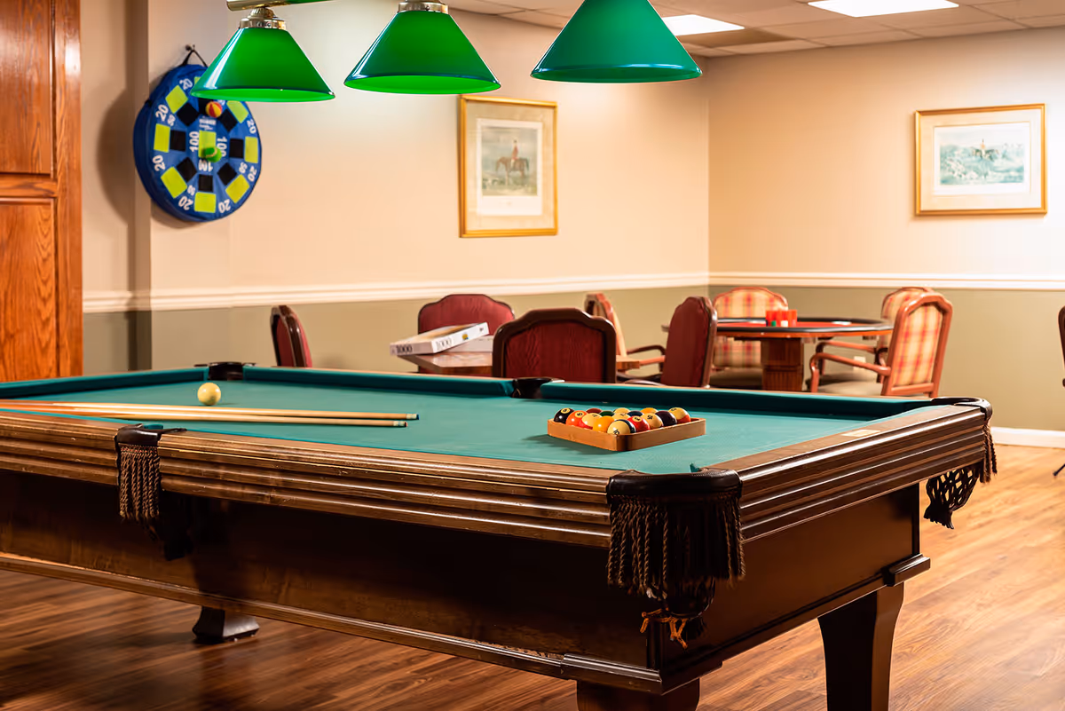 Pool table with green felt and hanging green lamps in a game room with tables and chairs in the background.