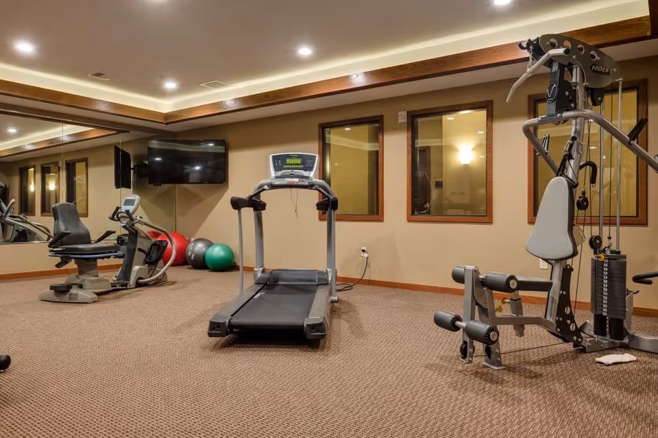 Interior view of a fitness room in a senior living facility featuring exercise equipment including a treadmill, stationary bike, multi-function weight machine, and exercise balls. The room has beige walls, carpeted floor, recessed lighting, a wall-mounted TV, and large mirrors on one side.
