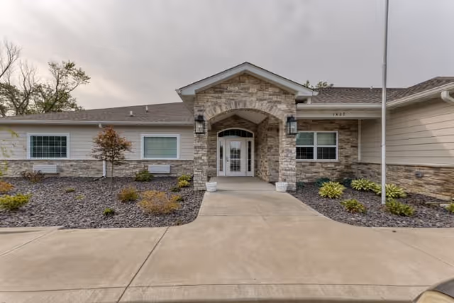 Front exterior view of a single-story building with a stone and siding facade, featuring a covered entrance with an arched stone design and glass double doors. There are small landscaped areas with shrubs and young trees on either side of the concrete walkway leading to the entrance.