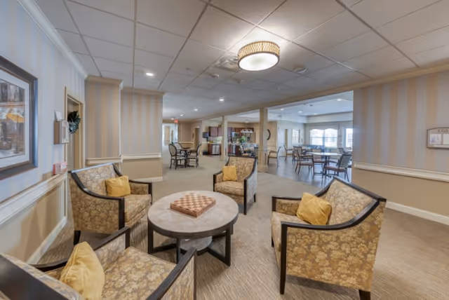 A spacious and well-lit common area in a senior living facility featuring four upholstered armchairs with yellow cushions arranged around a round table with a chessboard on top. In the background, there are additional tables and chairs, a kitchenette area, and large windows letting in natural light. The walls are painted in neutral tones with striped wallpaper accents and framed artwork.