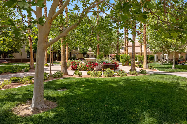 Shaded courtyard at a senior living facility with trees, green lawn, flowerbeds, benches, and a central fountain.