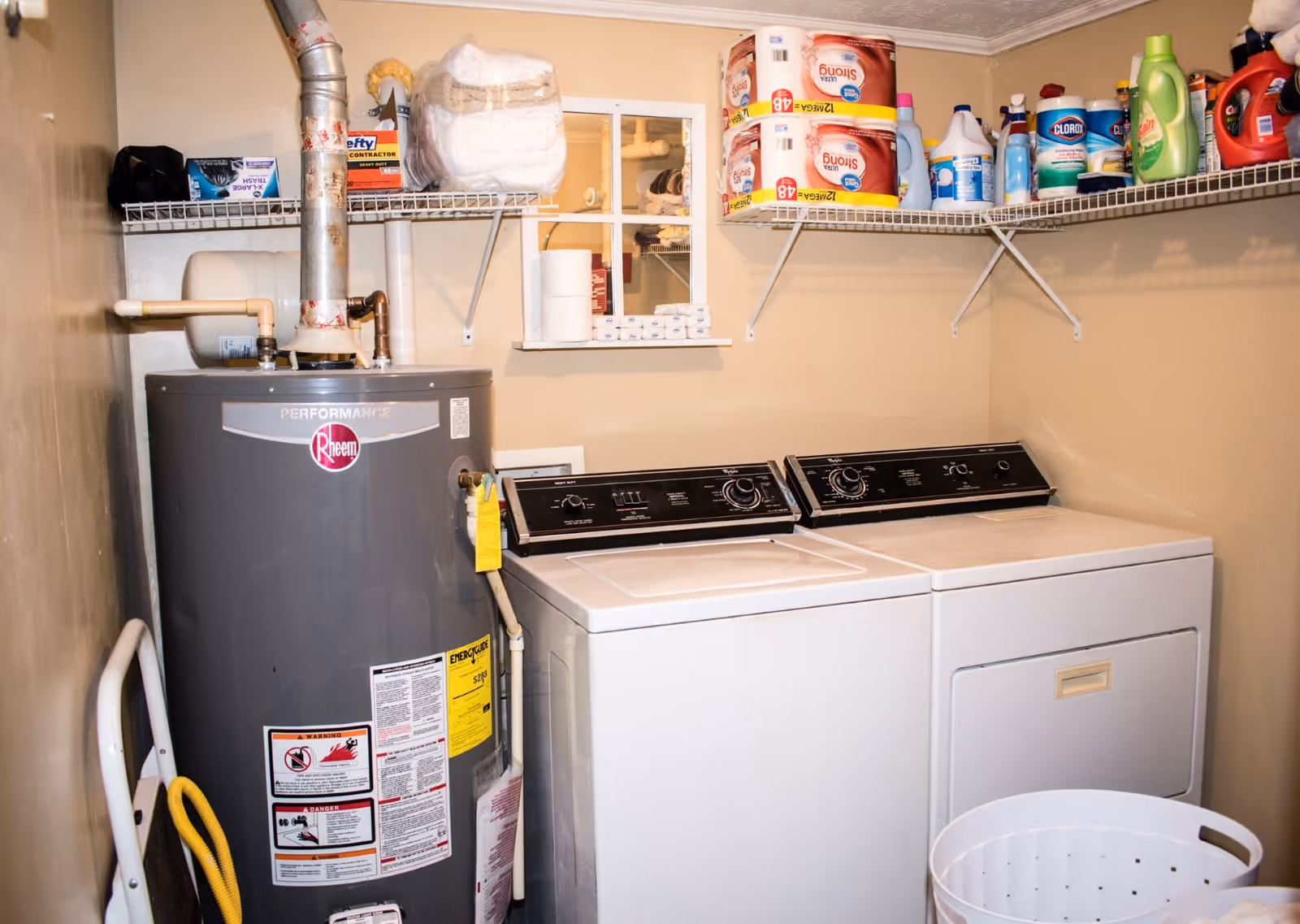 Laundry room with a Rheem water heater on the left, a washing machine and dryer side by side on the right, and shelves above holding various cleaning supplies, paper towels, and laundry detergent. A white laundry basket is partially visible in the foreground.