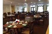 A spacious dining room with multiple round wooden tables set with plates, cups, and utensils. The room has several chairs around each table, framed pictures on the walls, and large windows with curtains allowing natural light to enter.