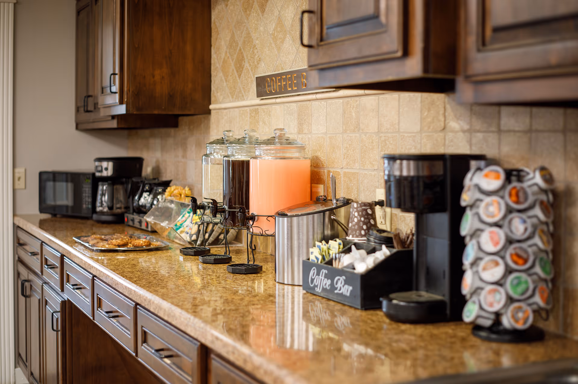 A kitchen counter with various beverage dispensers, a coffee maker, a microwave, and a rotating rack filled with coffee pods. The counter also has a tray with cookies and a container labeled 'Coffee Bar' holding cups and coffee supplies. The cabinets above and below the counter are wooden, and the backsplash is tiled.