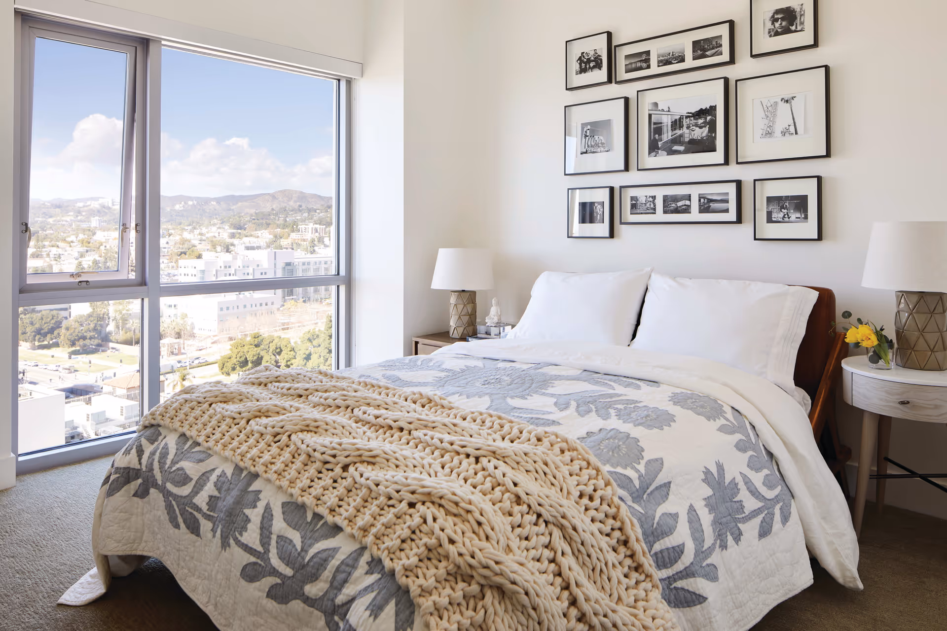 A bright bedroom with a large window showing a city and mountain view. The bed is neatly made with white pillows, a floral patterned quilt, and a chunky knit beige throw blanket. Two bedside tables each hold a lamp, and one has a small vase with yellow flowers. A gallery wall of black and white framed photos decorates the wall above the bed.