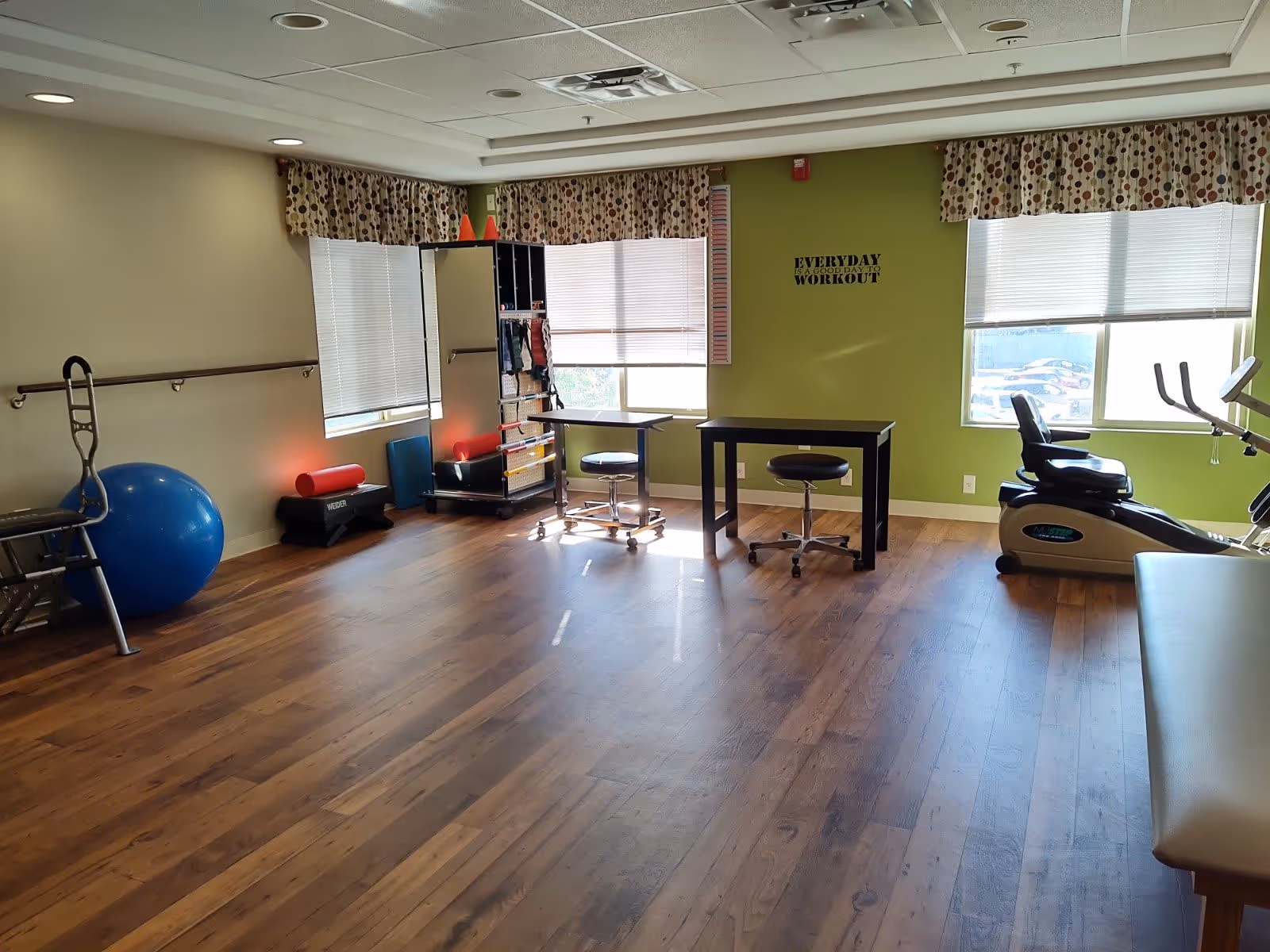 A bright exercise room with wooden floors and light green walls. The room contains various fitness equipment including a large blue exercise ball, a stationary bike, a mirror, and small tables with stools. Two windows with patterned valances allow natural light to enter. A wall decal reads 'EVERYDAY IS A GOOD DAY TO WORKOUT'.