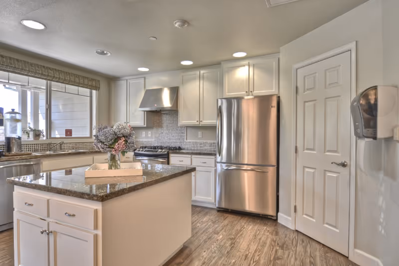 A modern kitchen with white cabinets, a stainless steel refrigerator and stove, a kitchen island with a granite countertop, and a vase of flowers on the island. There is a window with a beige valance above the sink, and wood-style flooring throughout.