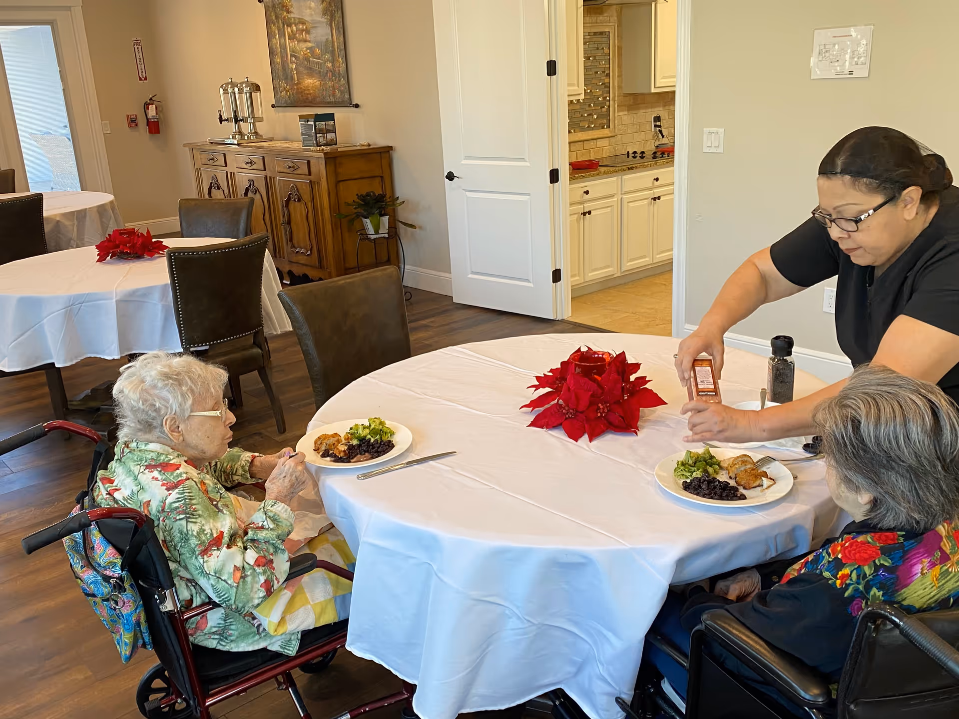 Two elderly women in wheelchairs seated at a round dining table with white tablecloths, each with a plate of food. A caregiver is seasoning the food of one of the women. The room has wooden flooring, additional tables with chairs, a wooden sideboard, and a kitchen visible through an open door.