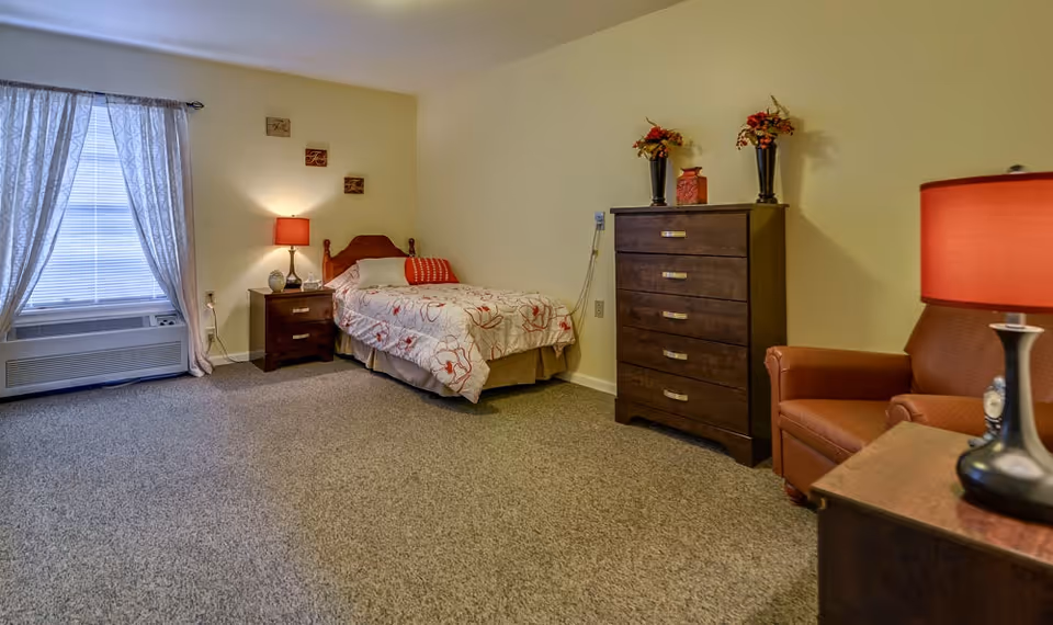 A senior living bedroom with a single bed covered in a floral bedspread, a wooden nightstand with a red lamp, a wooden chest of drawers with decorative vases and flowers on top, a brown armchair, and a window with sheer curtains letting in natural light.