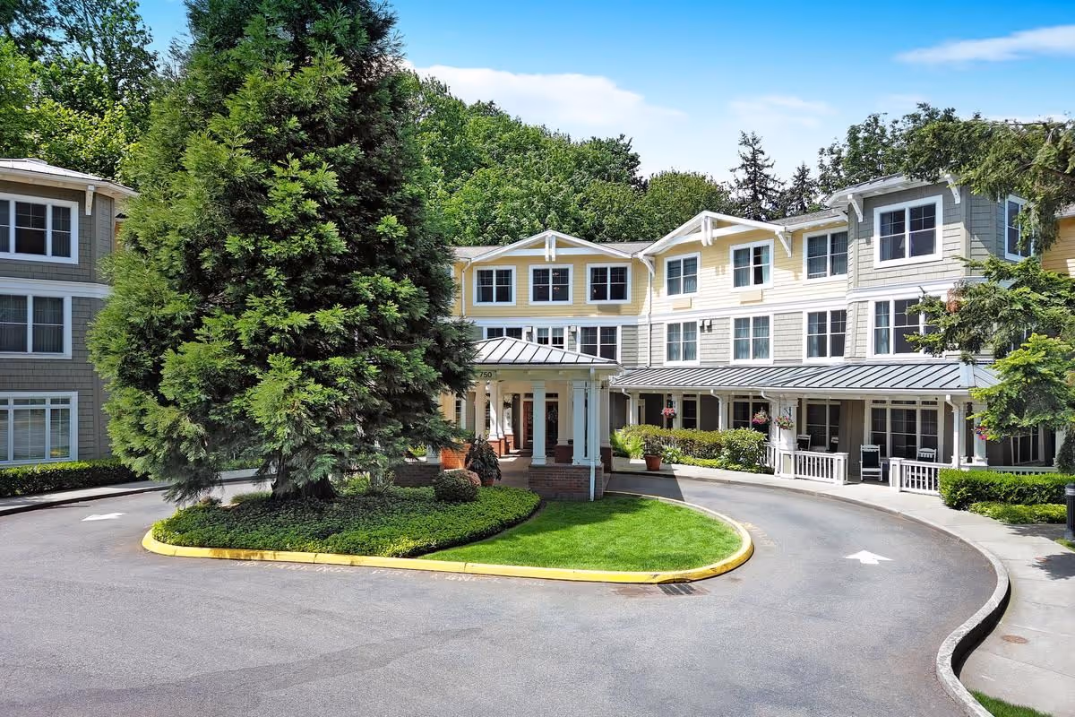 Front exterior view of a senior living facility named Sunrise of Edmonds, featuring a circular driveway with a landscaped island containing a large tree and greenery, surrounded by a three-story building with multiple windows and a covered entrance.