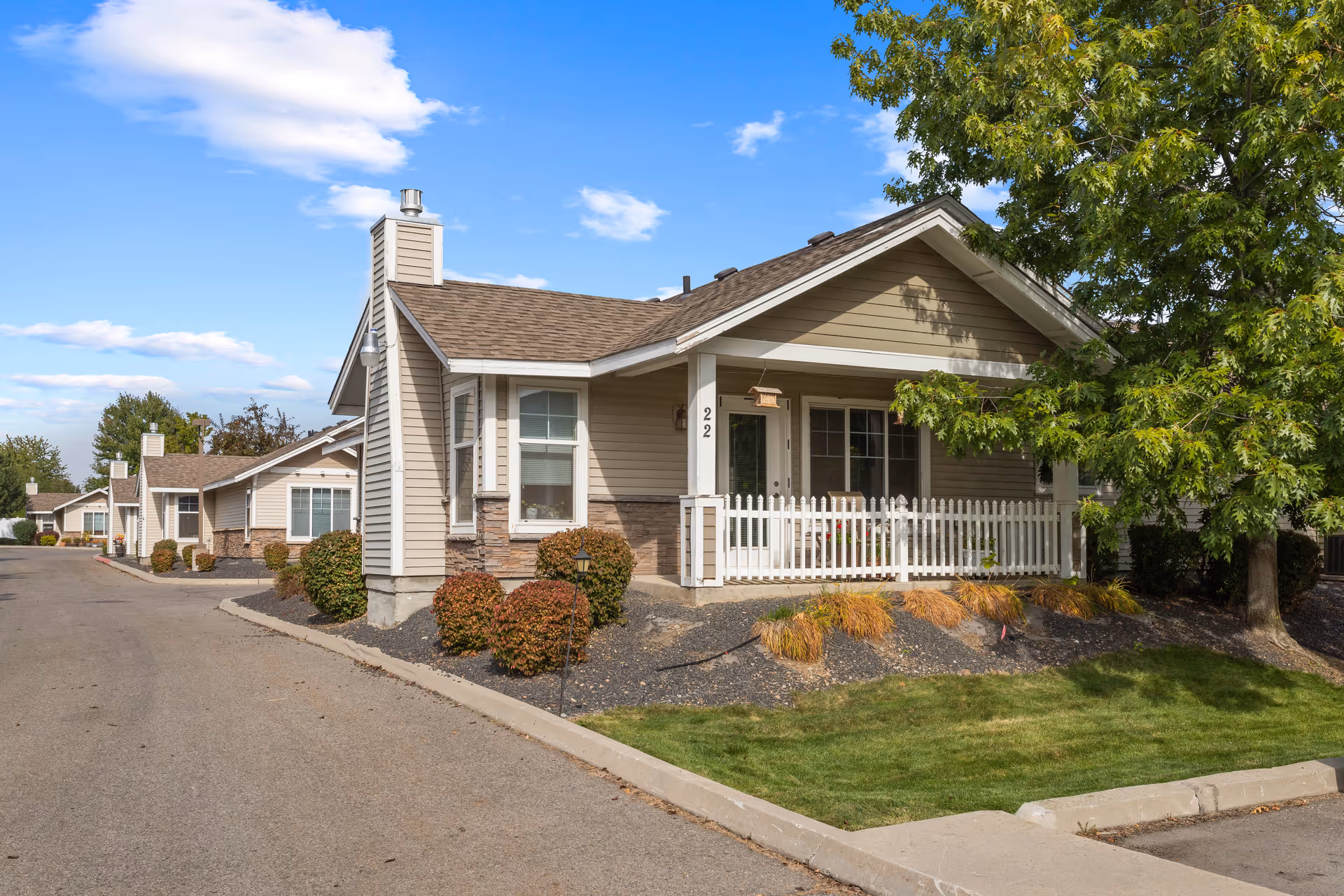 Exterior view of a single-story senior living cottage with beige siding, a small white picket fence porch, and a well-maintained lawn and shrubs under a blue sky with some clouds.