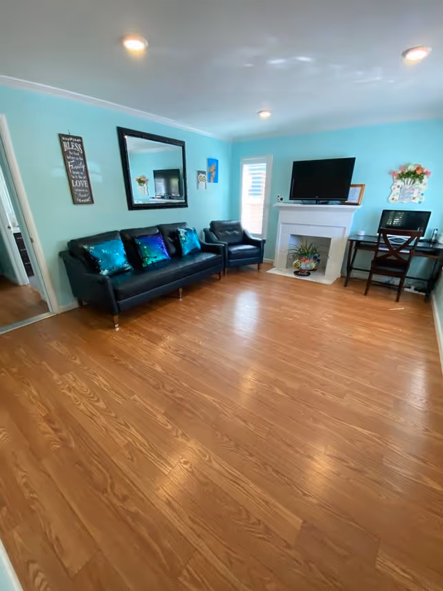 A living room with light blue walls and wooden flooring. The room features a black leather sofa with three shiny blue-green pillows, a matching black leather armchair, a white fireplace with a flat-screen TV on top, and a small desk with a chair and computer. There is a large rectangular mirror on the wall above the sofa, decorative wall art, and a potted plant inside the fireplace.