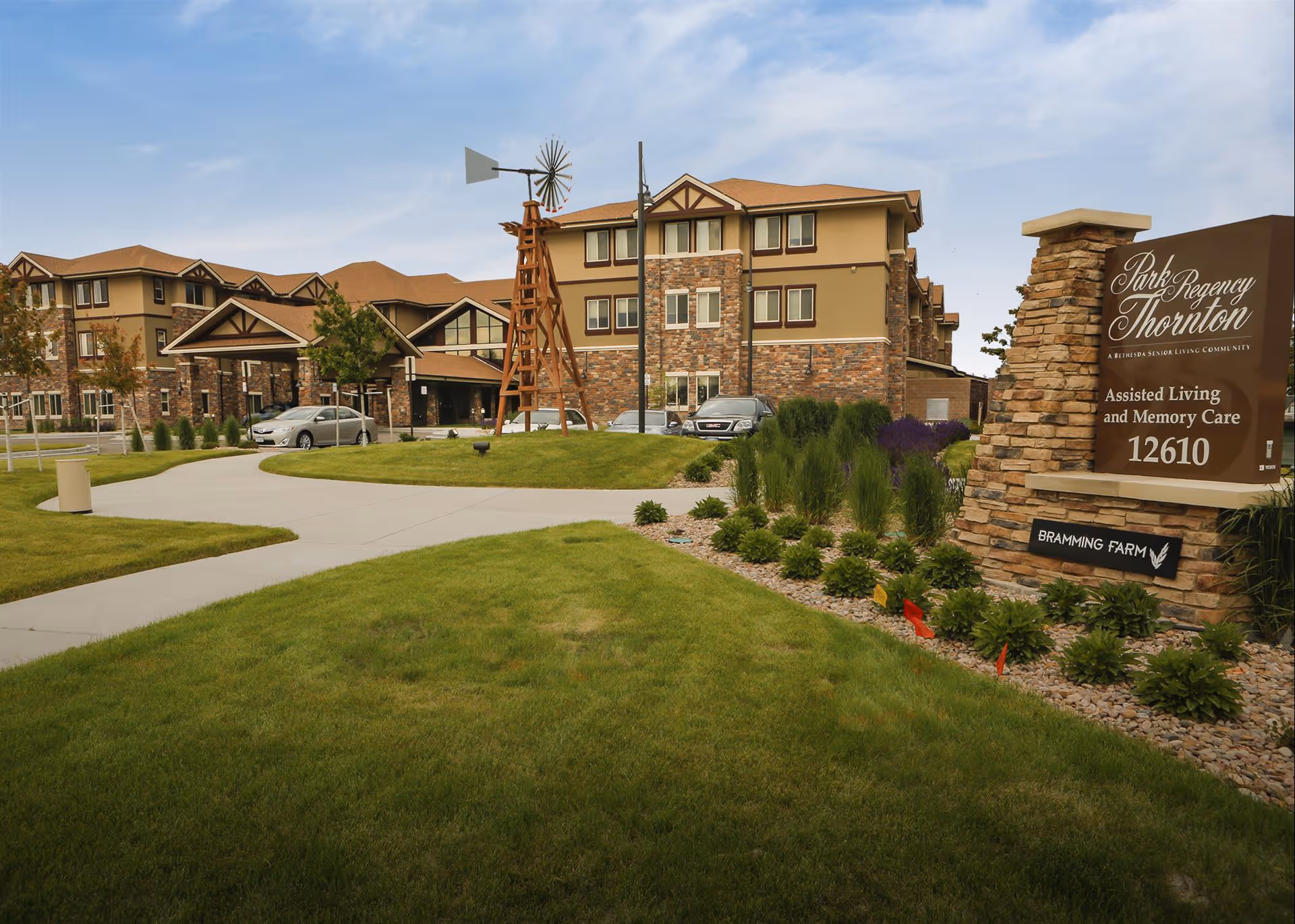 Exterior view of Park Regency Thornton, an assisted living and memory care facility. The building has a combination of stone and beige siding with a brown roof. There is a landscaped lawn with a concrete walkway, a wooden windmill structure, and a sign displaying the facility name and address 12610.