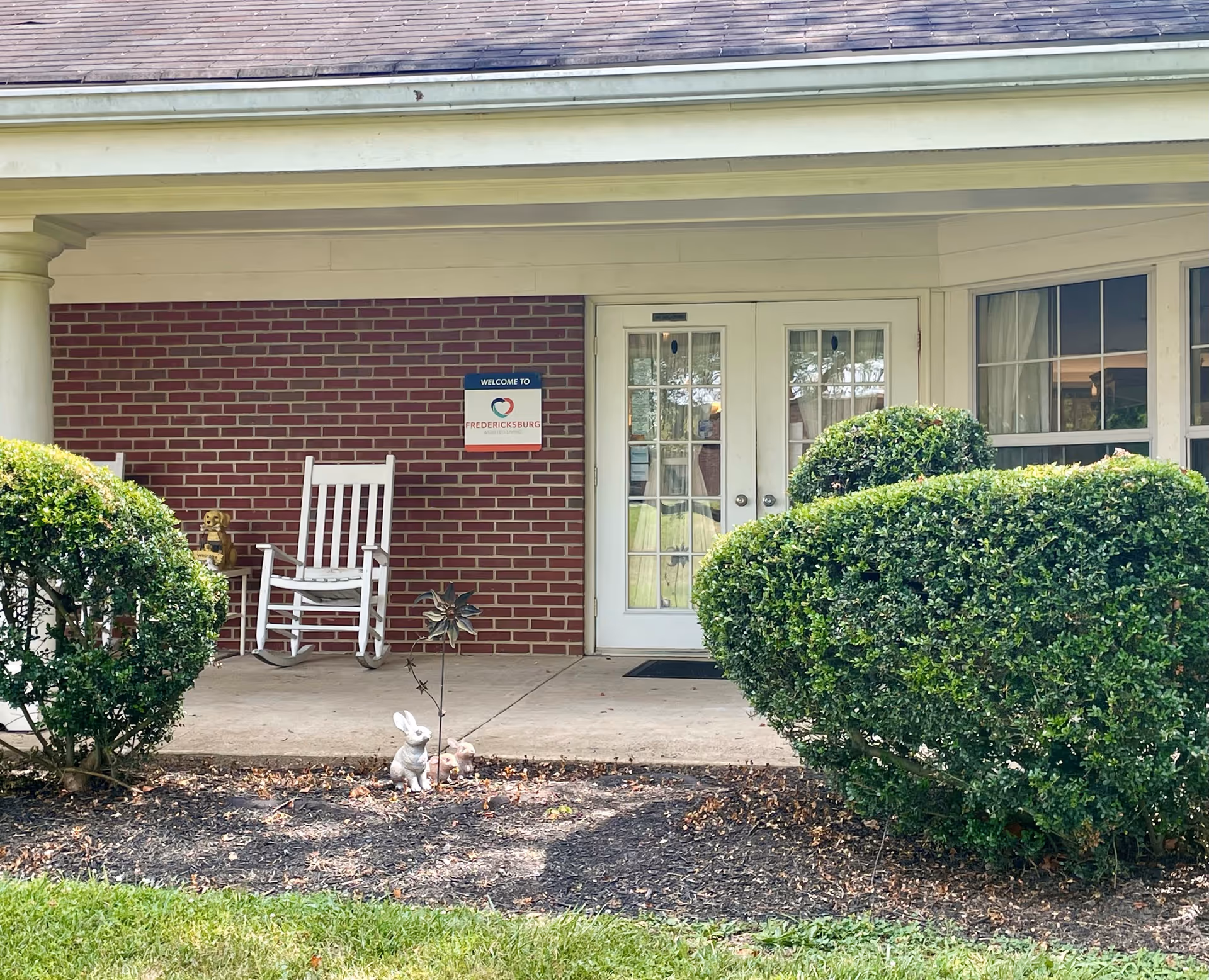 Front porch area of Fredericksburg Assisted Living featuring a white rocking chair, a small side table with a decorative item, a welcome sign on a red brick wall, double glass doors, and neatly trimmed bushes in the foreground.