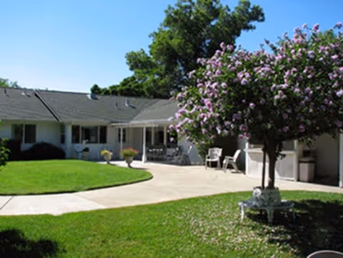 Outdoor courtyard area of a single-story assisted living facility with a well-maintained lawn, a flowering tree, patio chairs, and a covered seating area attached to the building under a clear blue sky.