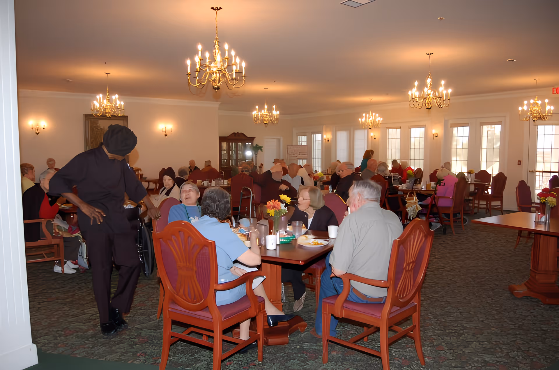 A large dining room in an assisted living facility with multiple elderly residents seated at tables eating and socializing. The room is well-lit with several chandeliers and wall sconces, and has large windows letting in natural light. A staff member dressed in dark clothing is walking among the tables. The furniture includes wooden chairs with red cushions and wooden tables with flower centerpieces.