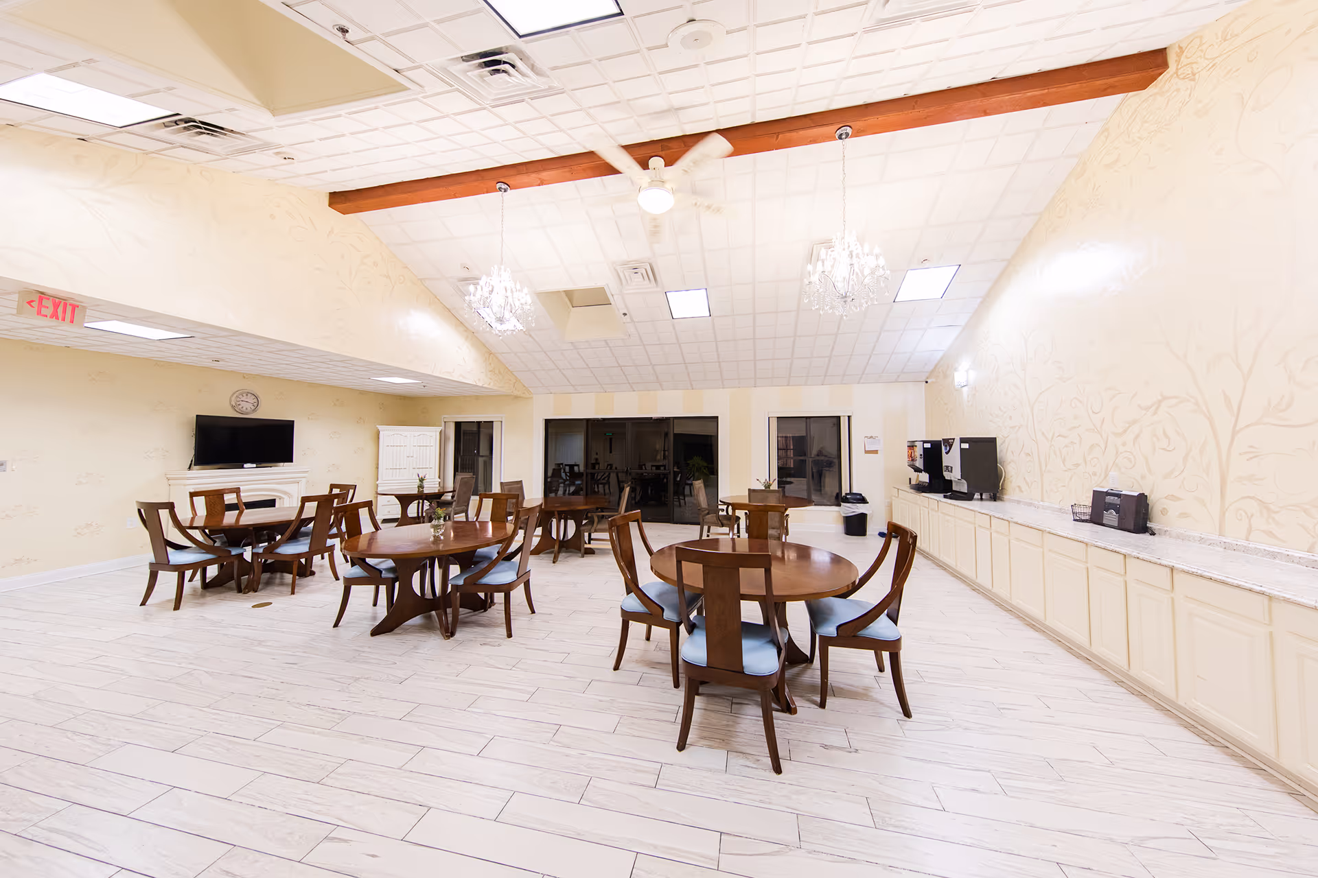 A spacious dining room with several round wooden tables and chairs with blue cushions. The room has a high ceiling with two chandeliers and a ceiling fan. There is a TV mounted on the wall above a white fireplace, a white cabinet, and a long counter with coffee machines and a trash bin. The walls are light-colored with subtle floral patterns, and the floor is covered with light wood-like tiles.