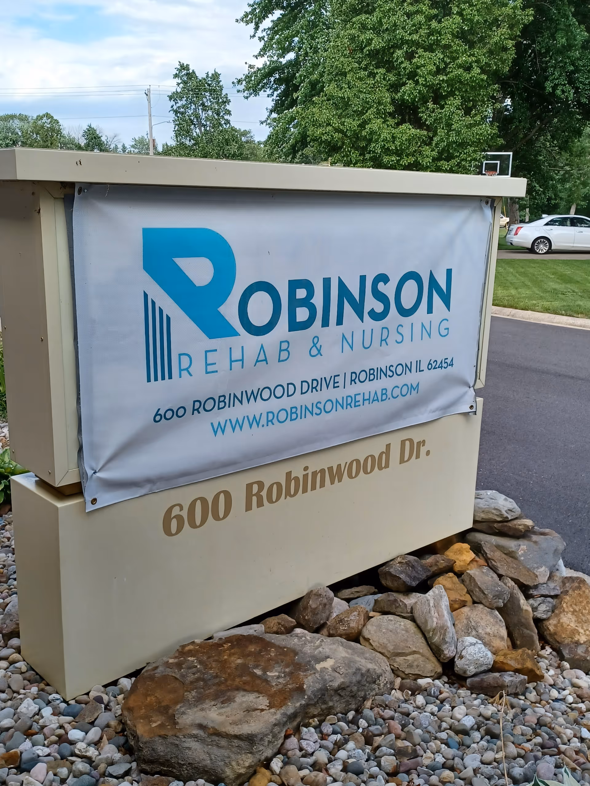 Outdoor sign for Robinson Rehab & Nursing located at 600 Robinwood Drive, Robinson, IL 62454, with the website www.robinsonrehab.com. The sign is surrounded by rocks and there is a road and grassy area with trees and a parked car in the background.