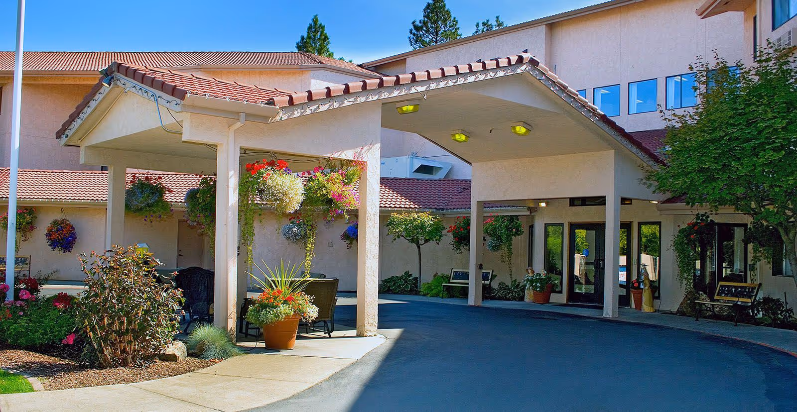 Covered front entrance of Solista Spokane with hanging flower baskets, potted plants and a driveway leading to the building's glass doors.
