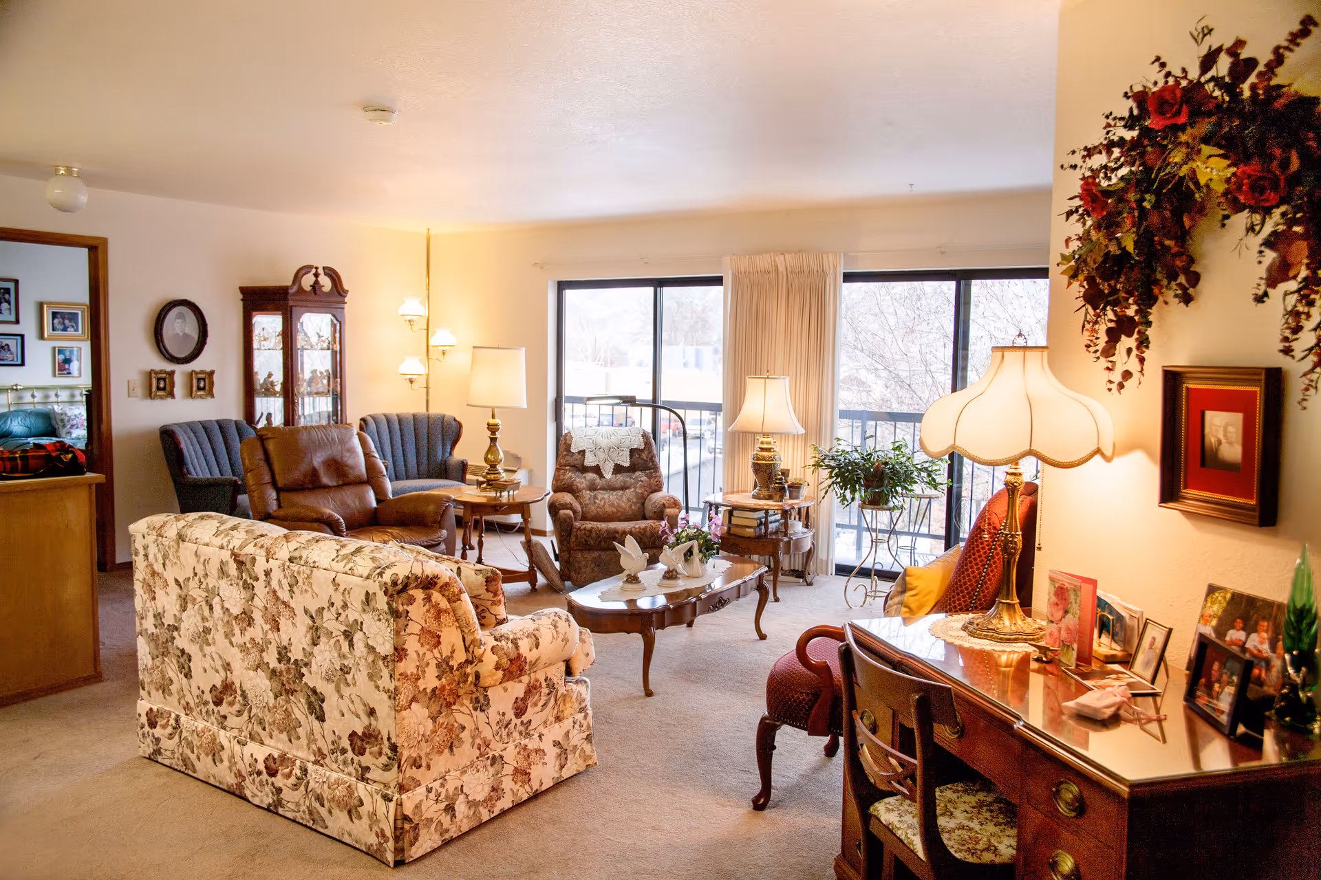 A cozy living room in Evergreen Estates featuring a floral-patterned sofa, a brown leather recliner, two blue armchairs, and a patterned armchair with a lace doily. The room has large windows with beige curtains letting in natural light, several table lamps, a wooden coffee table with decorative items, a wooden desk with framed photos and a lamp, and a floral wall decoration above the desk.