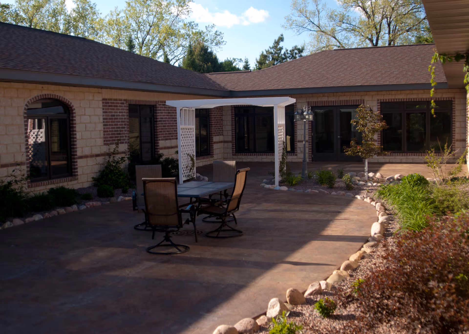 Sunlit courtyard patio with a table and chairs beneath a white pergola in front of a single-story brick building.