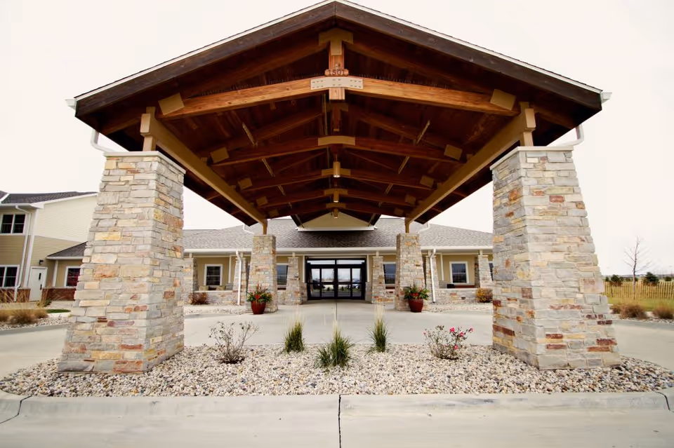 Entrance of The Village at Mercy Creek featuring a covered driveway with a wooden roof supported by stone pillars, landscaped with small plants and rocks, leading to glass doors of the building.